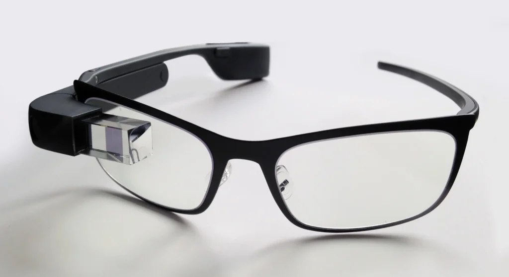 Google Glass: Too Early or Just Wrong? 2 A Google Glass smart device attached to a pair of black, rectangular prescription eyeglasses resting on a white surface.